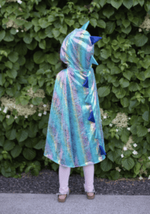 Child in colorful dinosaur cape by leafy wall.
