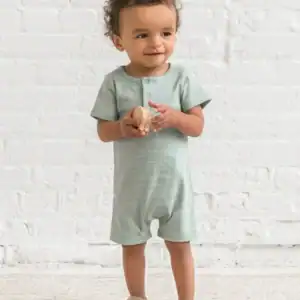 Toddler in romper holds wooden toy, smiling.