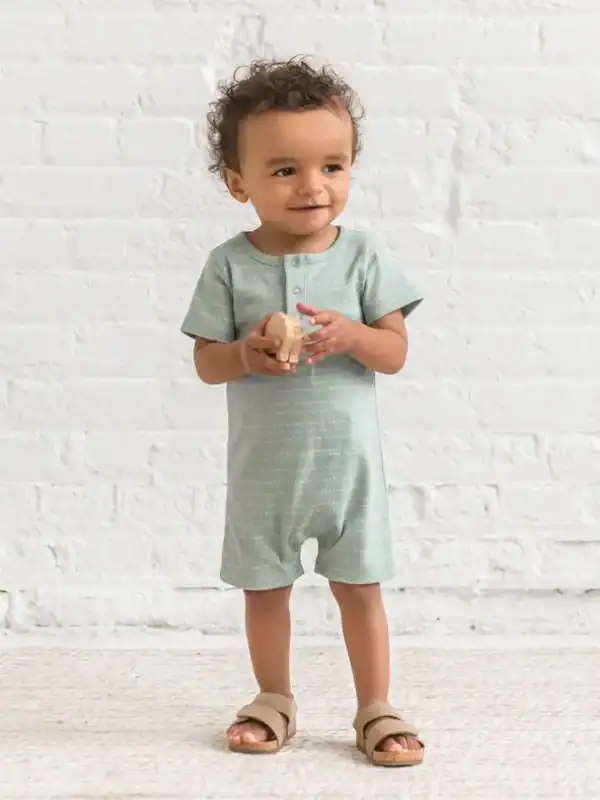 Toddler in romper holds wooden toy, smiling.