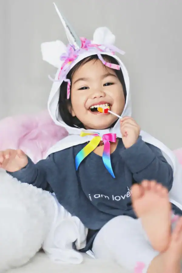 Child dressed as unicorn, smiling with lollipop.