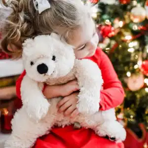 Child hugs teddy bear by Christmas tree.