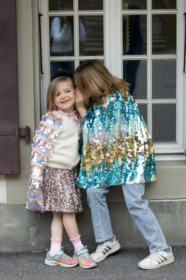 Two children whispering, wearing sparkling clothes.