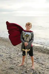 Child in knight costume with sword on beach.