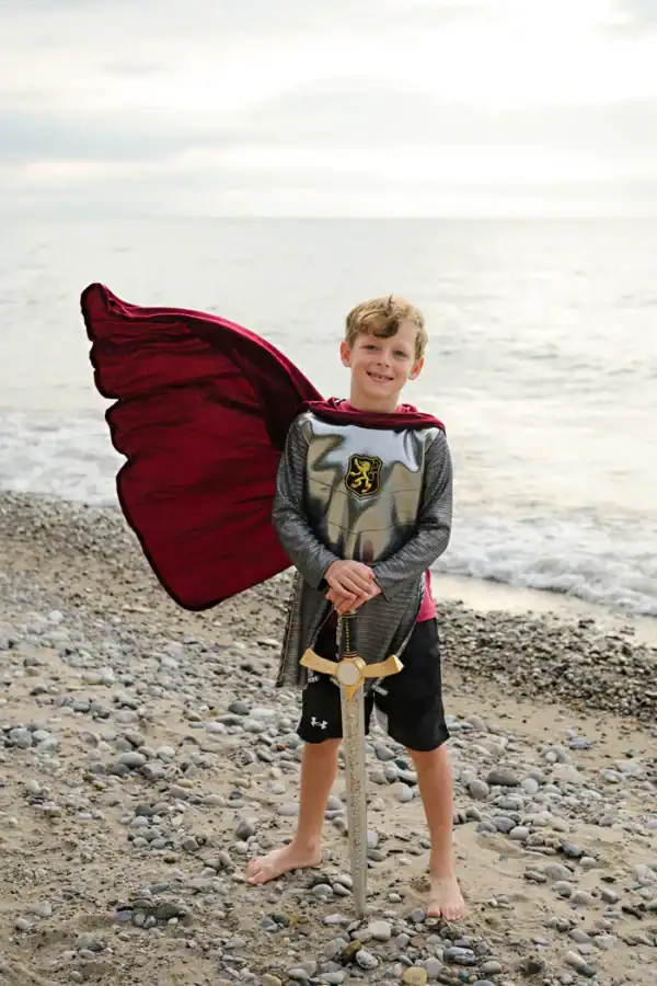 Child in knight costume with sword on beach.